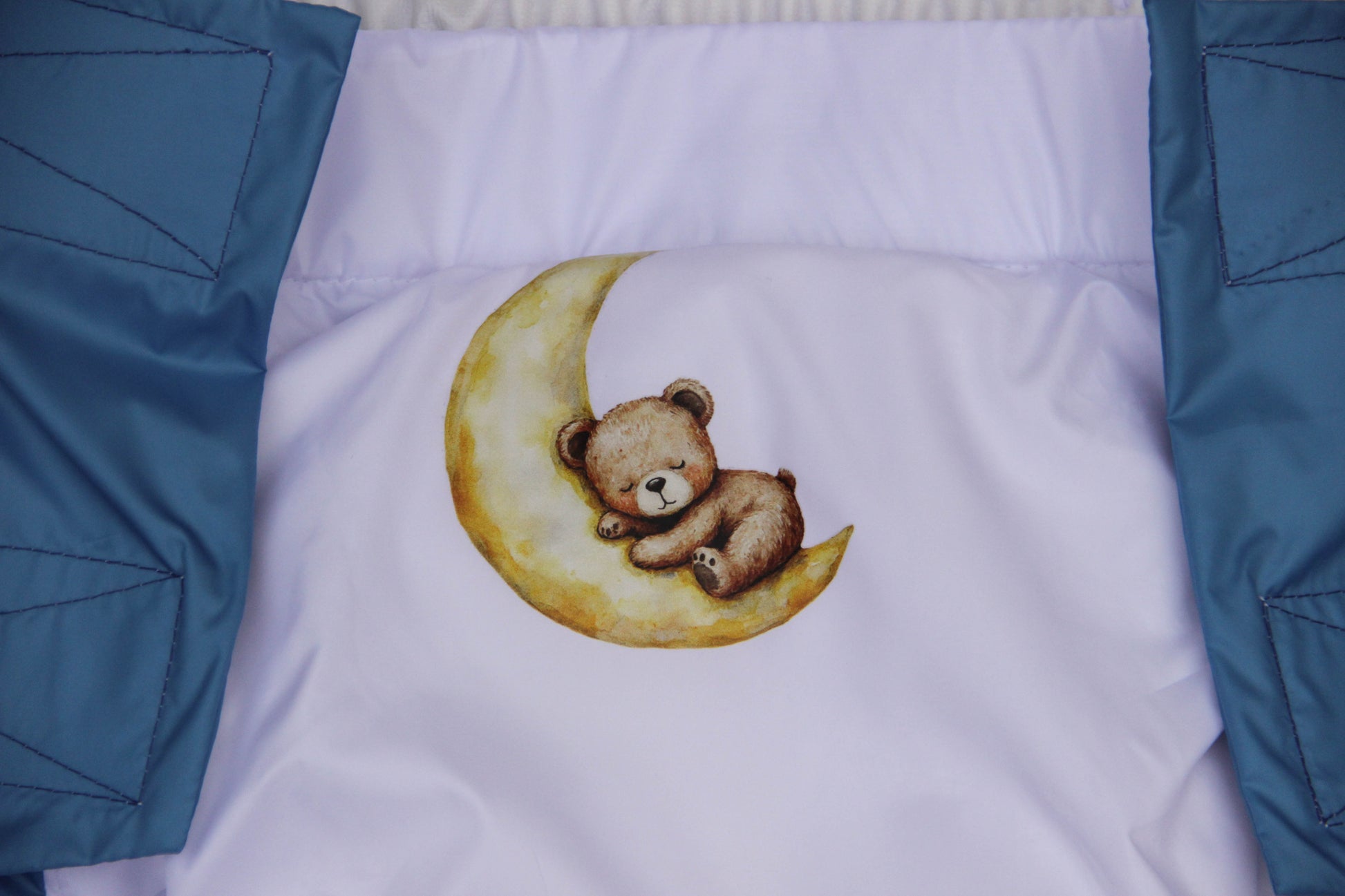 Embroidered teddy bear on a moon design on a white fabric with blue borders.